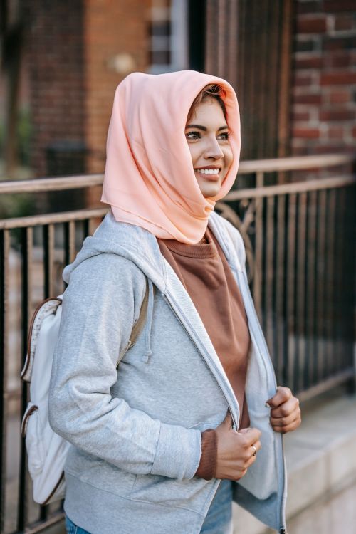 Free Side view of happy young Muslim female tourist in casual clothes and hijab with backpack smiling and looking away while standing on street near aged brick building Stock Photo