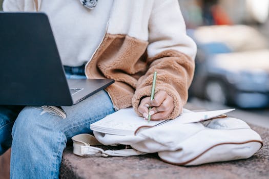 Woman sitting outside, using laptop and writing in notebook, working remotely in urban setting.