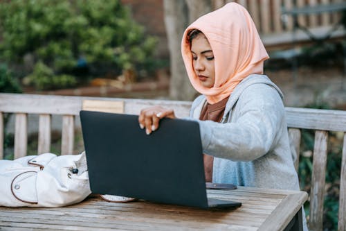 Concentrated Muslim woman wearing orange headscarf preparing portable laptop for remote work while sitting at wooden table in park in daylight
