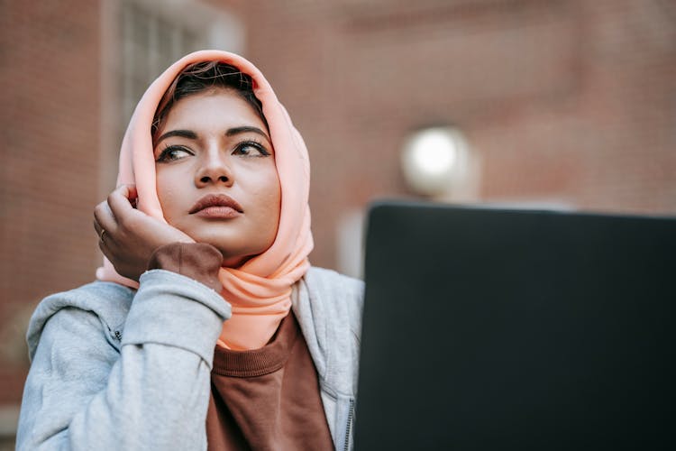 Muslim Female Student Daydreaming While Sitting With Laptop Near City Building