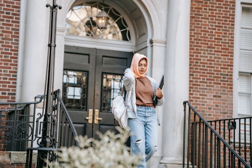 Young Muslim woman wearing jeans and orange hijab carrying backpack and laptop walking down stairway after studies in university