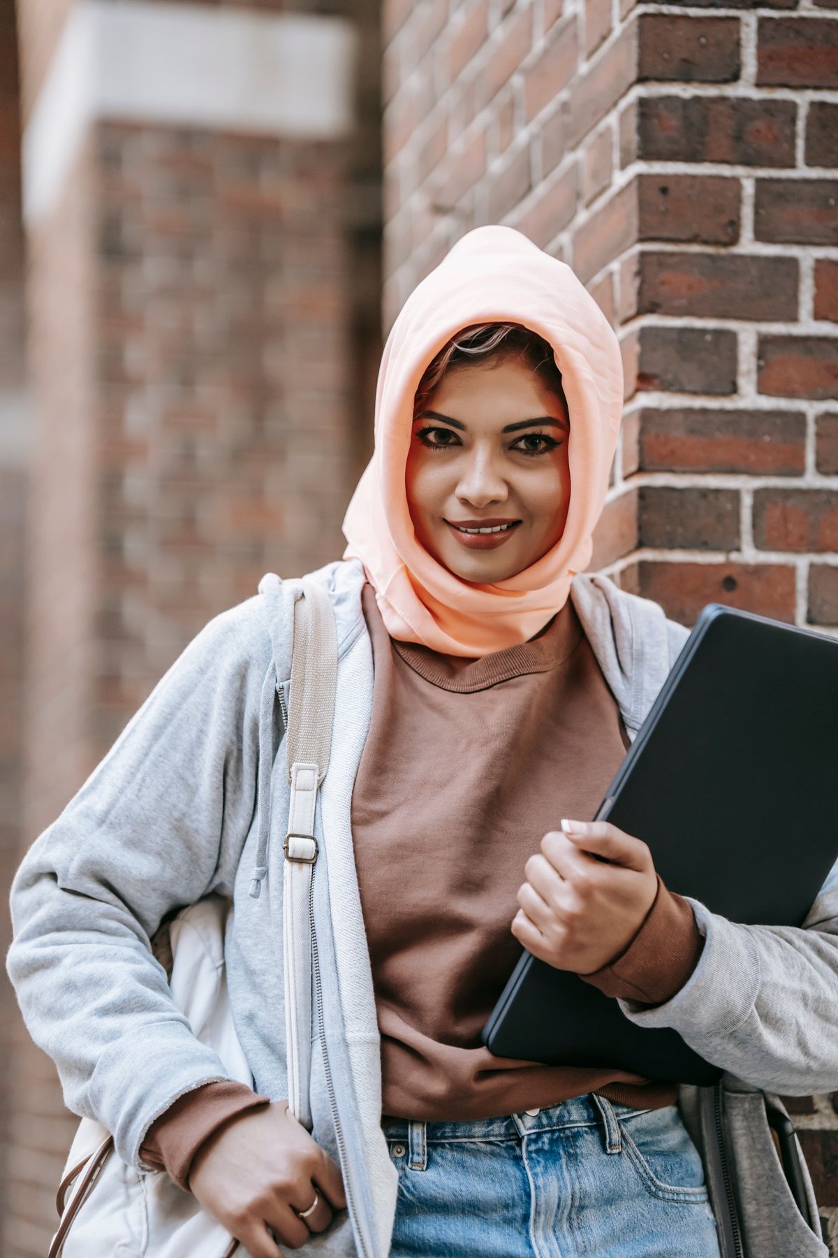 A confident student holding a laptop beside a brick campus building