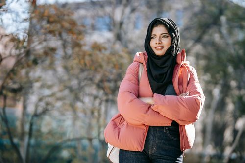 Free Smiling young Muslim female in pink jacket and traditional Islamic headscarf standing with hands crossed on street and looking away Stock Photo