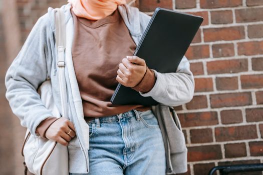 Unrecognizable ethnic woman in casual wear with bag and laptop standing beside brick wall on street