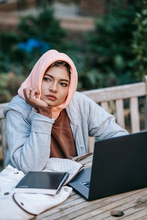 High angle of young thoughtful Muslim female freelancer in casual wear and traditional headscarf sitting at table with laptop in garden