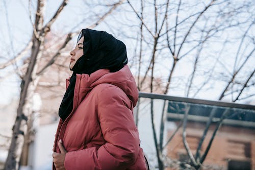 Low angle of young Muslim female in pink jacket and traditional black headscarf standing on street and looking away
