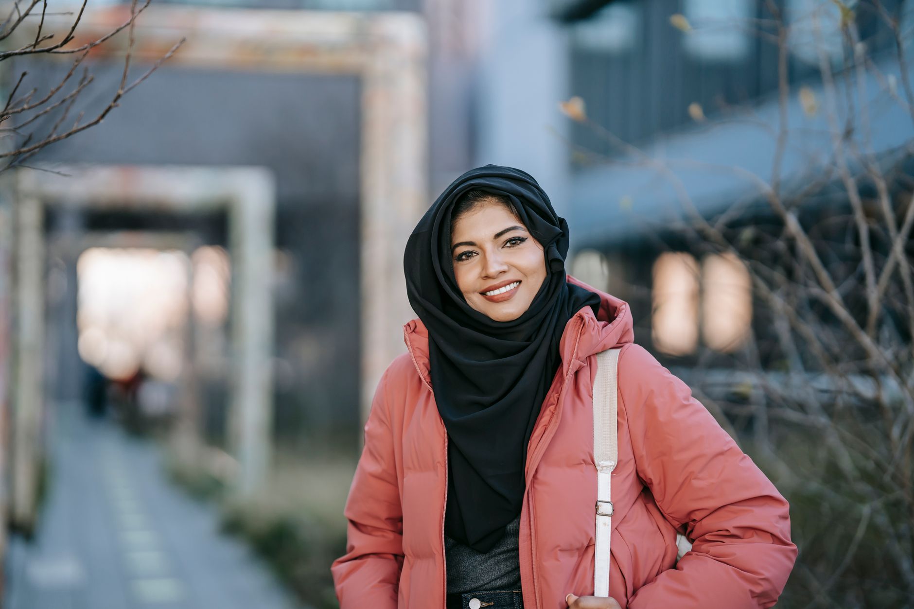 Happy Middle East woman in casual outfit and traditional Islamic headdress looking at camera in city street