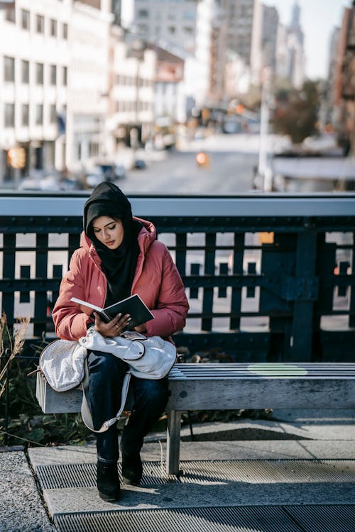 Free Full body of young Muslim female in casual clothes and black Islamic headdress sitting on bench and reading book on city street Stock Photo