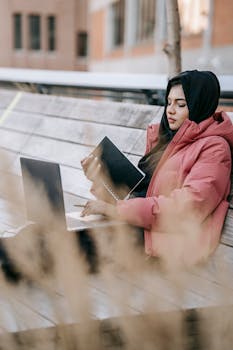 Focused woman in hijab using laptop on a bench, working remotely.