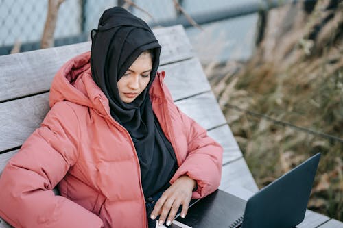 Confident young lady in warm outfit and hijab sitting on wooden bench while surfing on computer and notepad in daytime