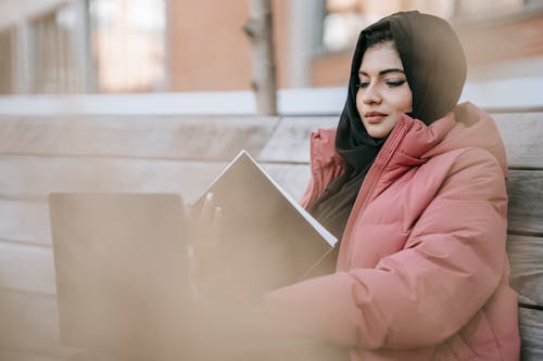 Free Confident young lady in hijab and warm outfit sitting on wooden bench while studying notepad and netbook in daytime Stock Photo