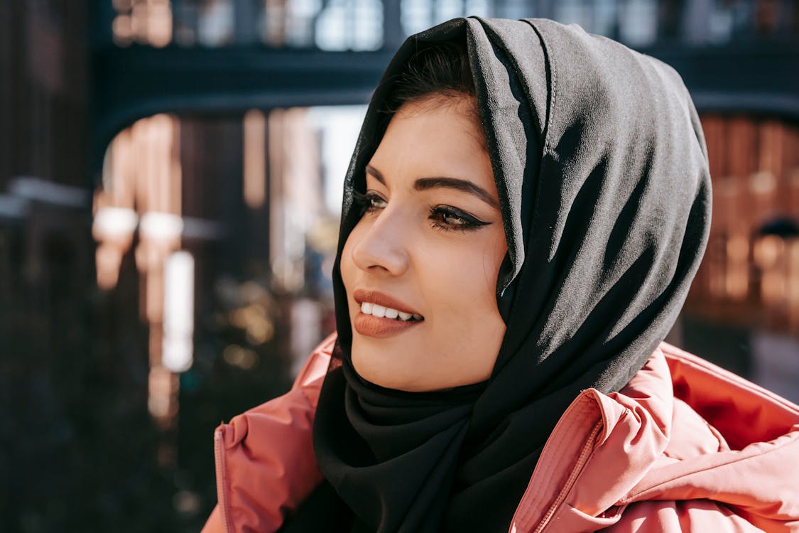 Content ethnic female in hijab smiling and looking away on blurred background of street