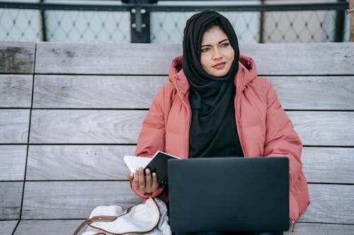 Thoughtful ethnic female in hijab with notebook in hand browsing laptop while resting on bench on street