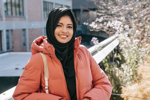 Free Cheerful ethnic female in hijab and jacket smiling and looking at camera on street Stock Photo