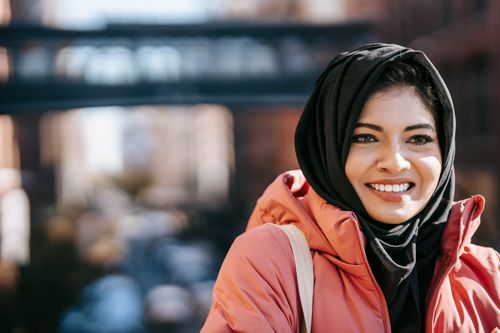 Free Muslim cheerful ethnic female in hijab smiling and looking away on blurred background of urban street Stock Photo