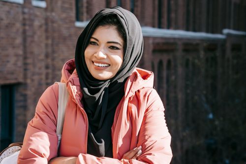 Free Cheerful ethnic female in hijab and jacket looking away on blurred background of brick building Stock Photo