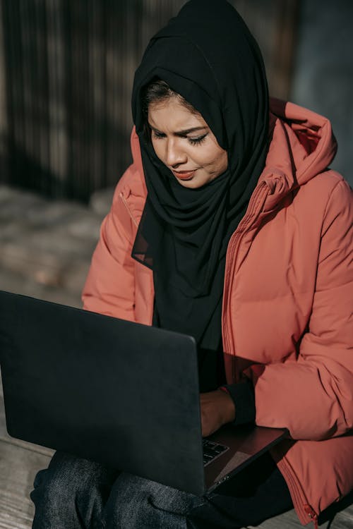 Thoughtful Muslim woman freelancer in casual clothes and headscarf sitting with laptop on knees while working on street in daytime