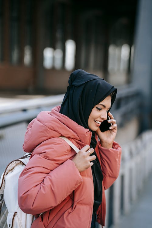 Cheerful Muslim female in casual clothes and black headscarf with backpack standing on street and talking on  smartphone on street in daytime