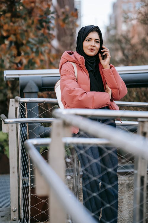 Calm Muslim female standing near metal fence and talking on smartphone