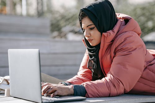 Pensive Muslim female in casual clothes and headscarf typing on netbook while working on project on street in daytime