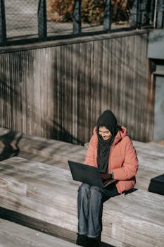 A smiling Muslim woman in a headscarf works on a laptop outdoors on a sunny day.