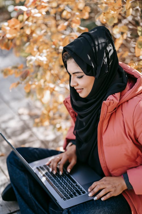 From above of focused Muslim woman freelancer in casual outfit and black headscarf sitting with netbook on knees while working on project on street against autumn tree in daytime