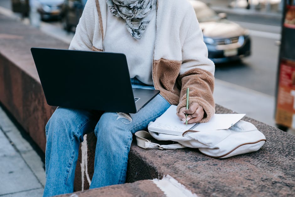 Does QuickBooks Self-Employed Help Track Write-Offs? Unrecognizable woman multitasking outdoors with laptop and notebook on a city bench.