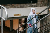 Charismatic woman in casual wear and headscarf with notebook in hand rising on stairs from underpass and looking at camera while having walk in city