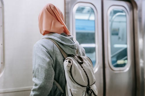 Back view of faceless Muslim woman in casual outfit with backpack and hijab standing near train in metro station