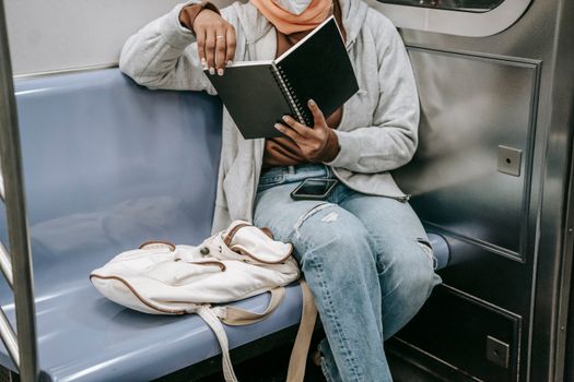 Faceless ethnic female sitting with notebook in train in mask