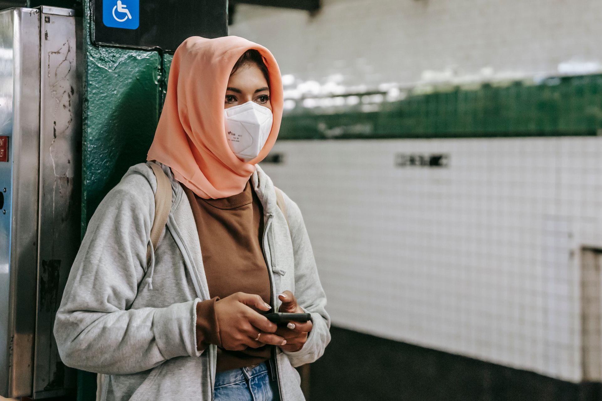 Faceless young Muslim ethnic woman in casual outfit with protective mask and hijab leaning on column in metro station while surfing on smartphone and looking away while waiting for train