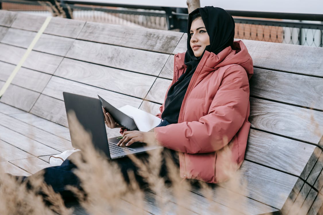 Young woman in warm clothes and hijab sitting on wooden bench in park and using laptop with notebook in daylight and looking away