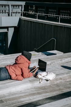 High angle of female freelancer in casual clothes lying on wooden roof while working on project using laptop in sunny autumn day