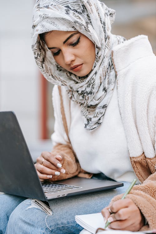 Free Young ethnic female freelancer in warm outfit and hijab using laptop while making notes in notebook with pen in daytime Stock Photo