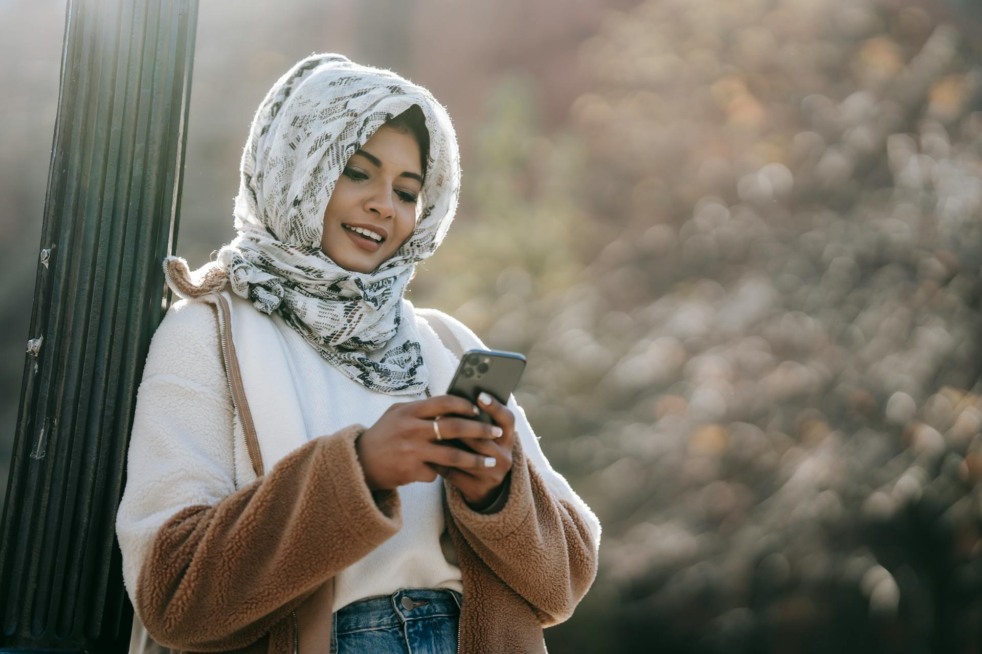 Smiling ethnic young woman in warm outfit and hijab leaning on pillar while text messaging on cellphone in daytime in city street