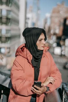 Serious ethnic young woman in hijab and warm outfit standing in city street near buildings while using smartphone in sunny day and looking away