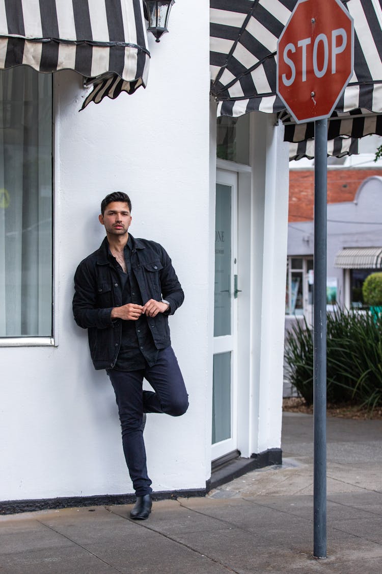 Man In Black Jacket Standing On One Leg Beside White Wall