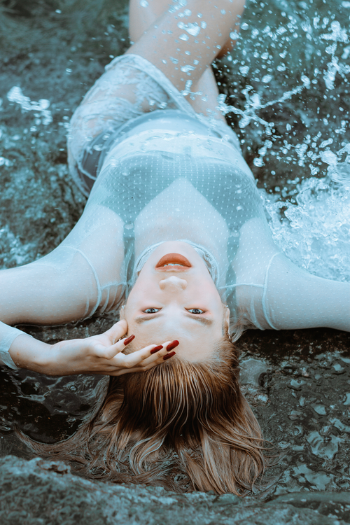 Free From above of young sensual female model in transparent cloth touching face while lying on wet sand in sea waves splashes Stock Photo