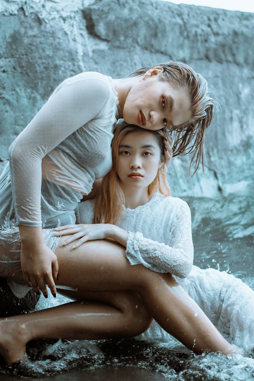 Calm barefoot female in transparent cloth sitting on stone and hugging Asian girlfriend lying in waving seawater against rocky cliff and looking at camera
