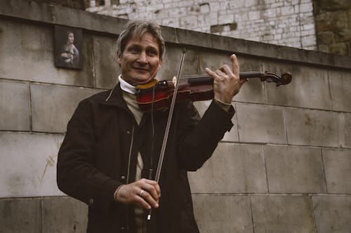 Kostenlos Lächelnder Stehender Mann, Der Geige Durch Graue Steinmauer Spielt Stock-Foto