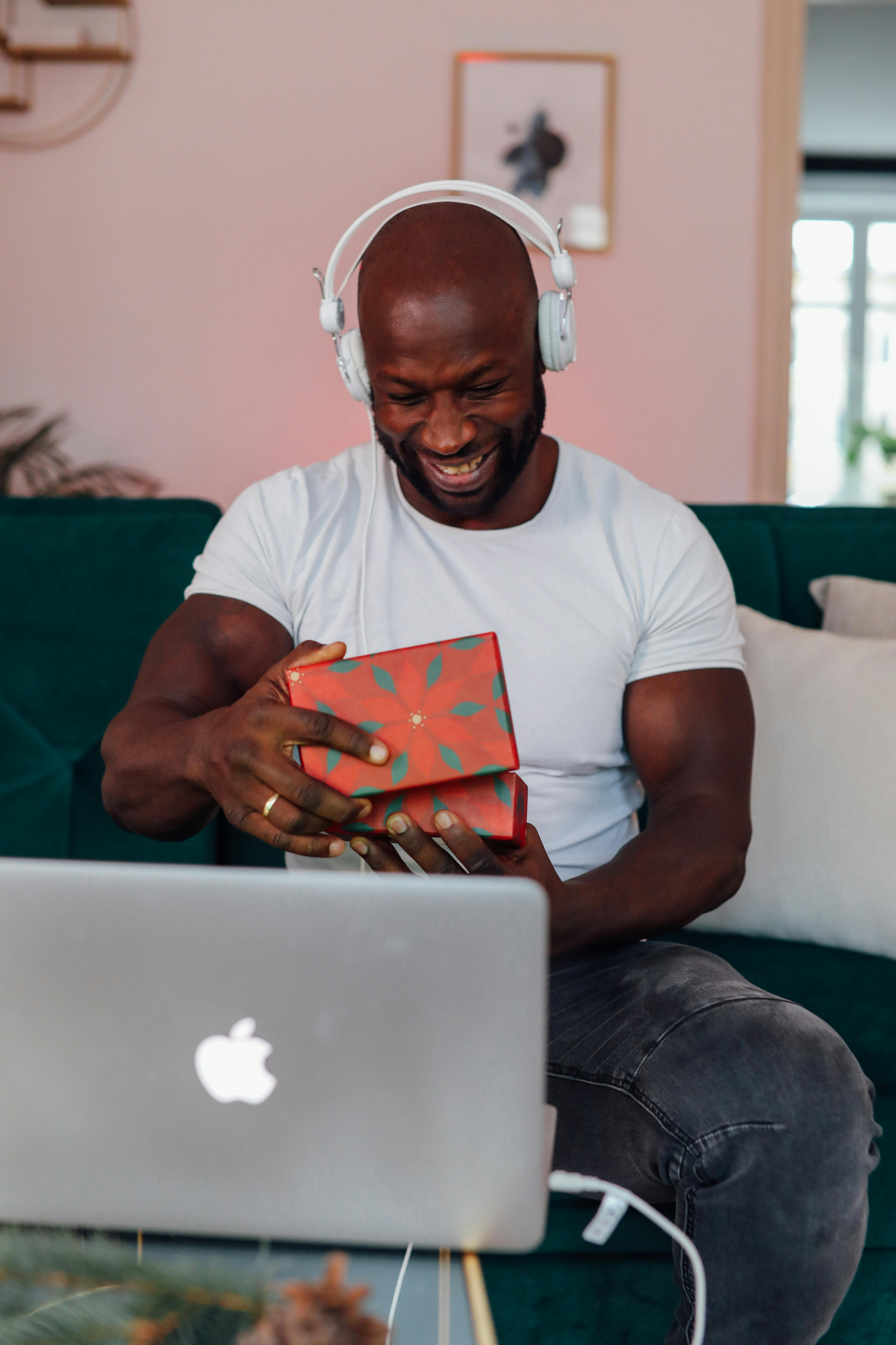 A joyful man wearing headphones opens a Christmas gift while sitting indoors with a laptop.