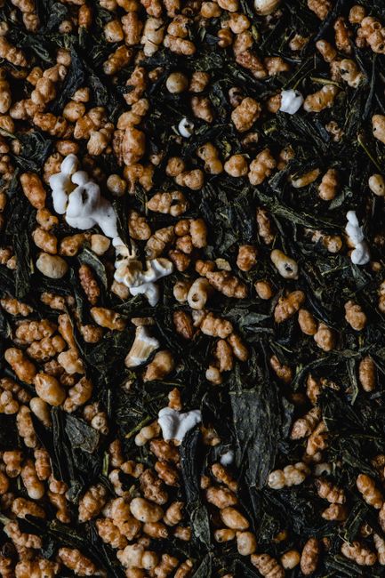 A glass jar of genmaicha showing dark green tea leaves mixed with pale roasted rice kernels, some popped open like tiny popcorn.
