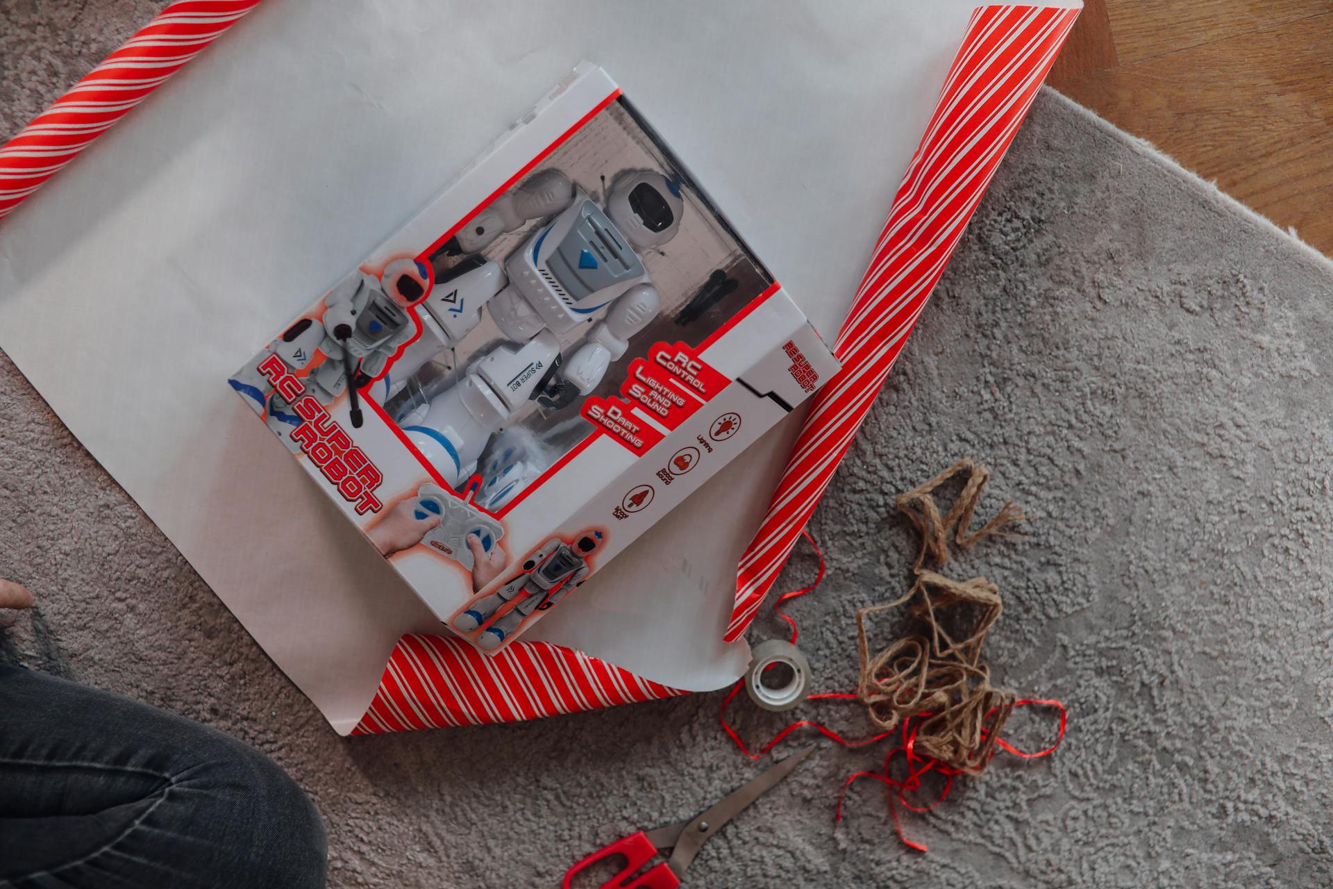 A remote control robot toy being wrapped as a Christmas present on a cozy carpet.