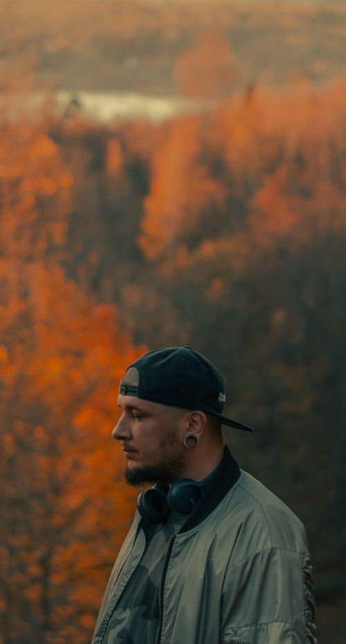 Free Side view of bearded male in cap with headphones looking away against lush trees in fall season Stock Photo