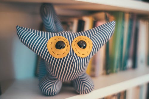 Adorable stuffed toy made of striped fabric placed on bookshelf with abundance of books in light room on blurred background