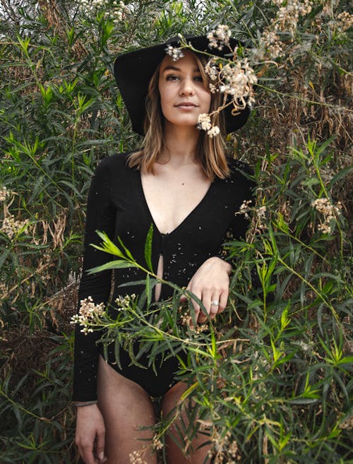Confident lady in black body with long sleeve and hat standing among tall green plants and looking at camera in summer day