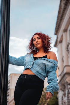 Fashionable woman in denim jacket posing outdoors with city architecture backdrop.