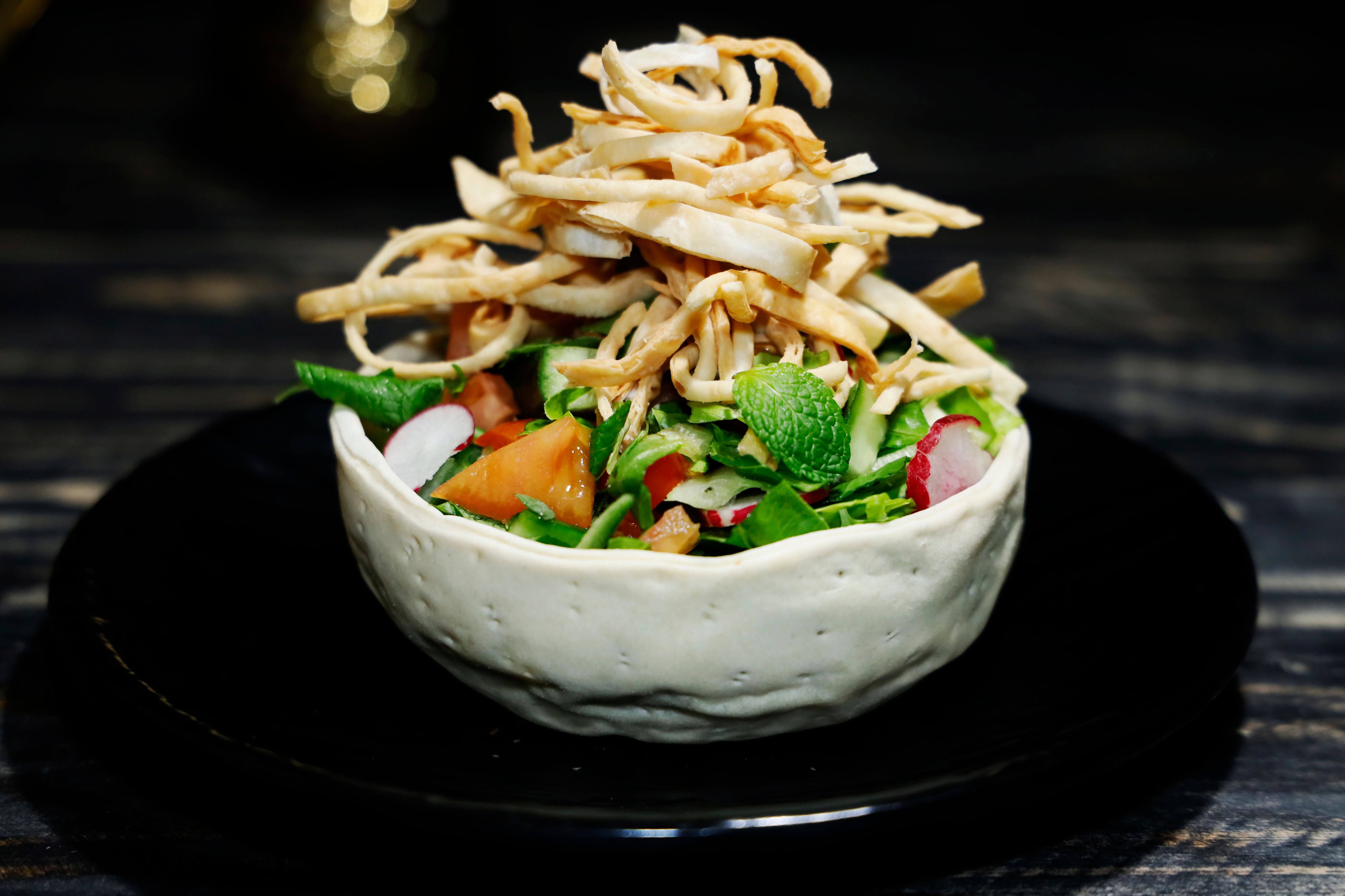 A vibrant Arabic salad topped with crispy fried strips served in a white bowl on a dark wooden table.