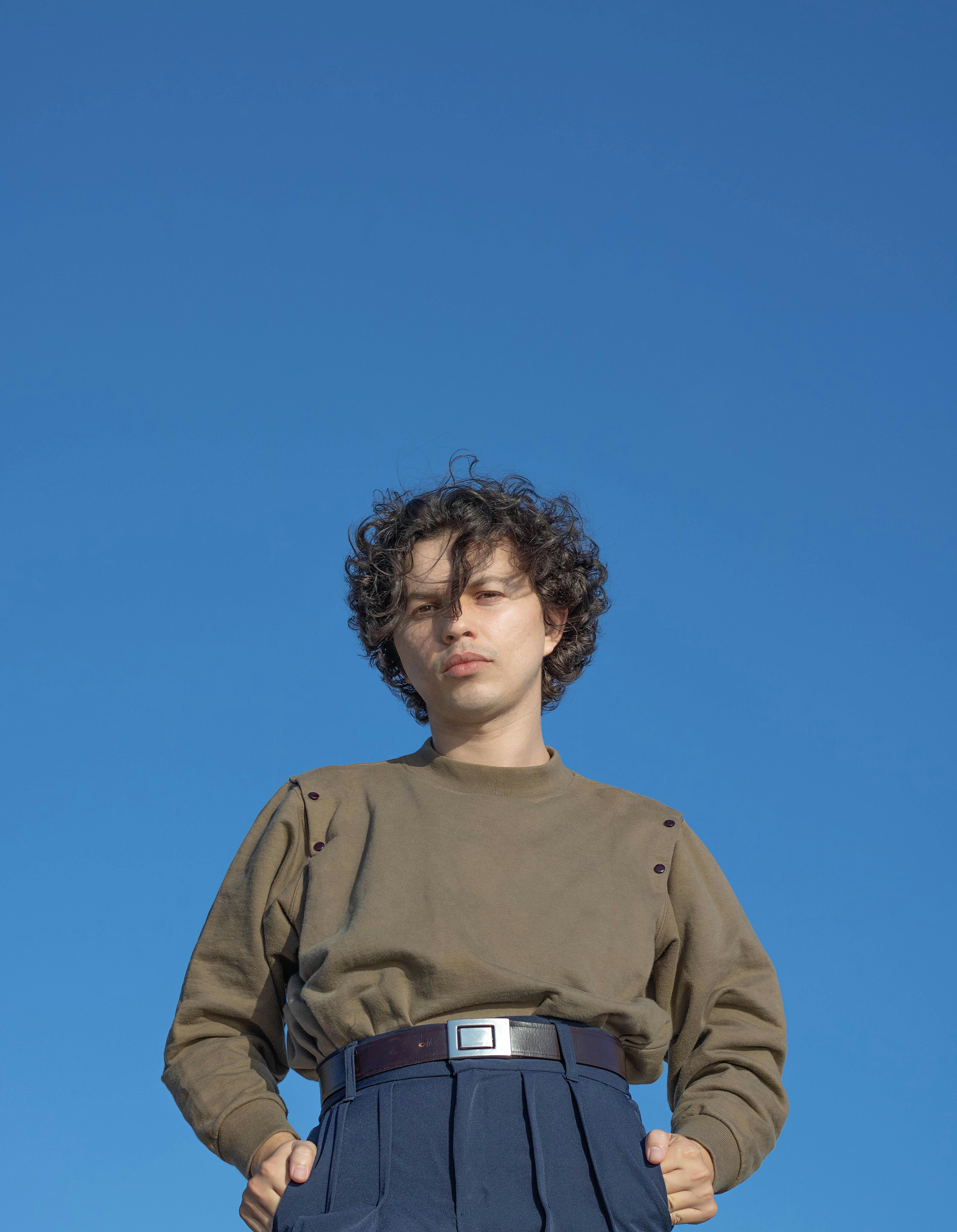 Low angle of cool millennial guy in trendy outfit looking at camera while standing on blue background