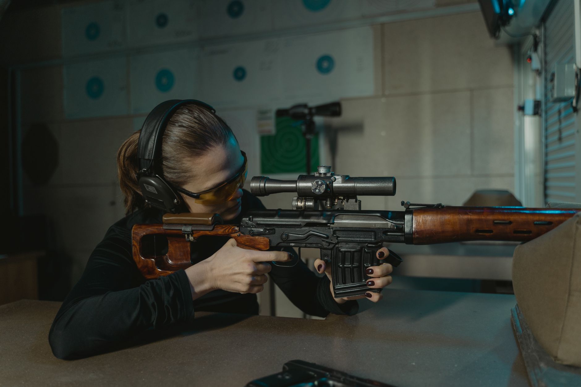 A woman with protective gear aiming a sniper rifle at an indoor shooting range.